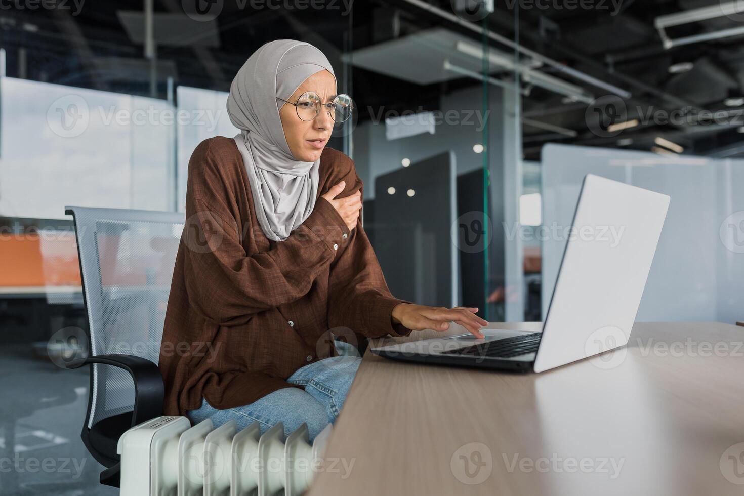 Businesswoman freezing in office at work, woman in hijab near heater trying to keep warm working inside office building with laptop, cold in office. photo