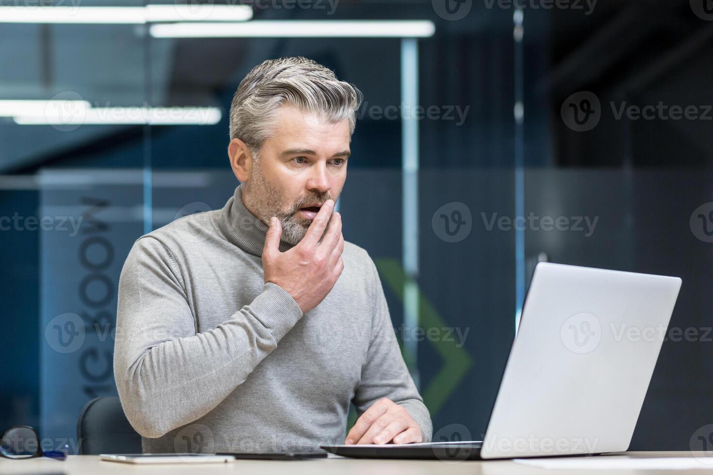 A shocked man, a gray-haired businessman, sits at the desk in the office, works with a laptop. He holds his hand to his mouth, surprised by what he saw on the computer screen. photo