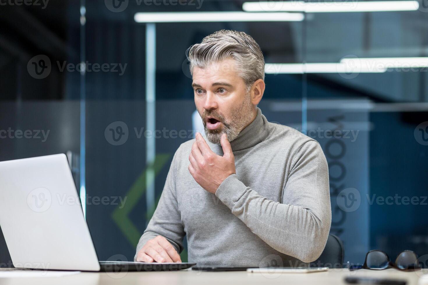 Senior gray haired businessman shocked reading bad news surprised and upset mature experienced man working with laptop inside modern office building. photo