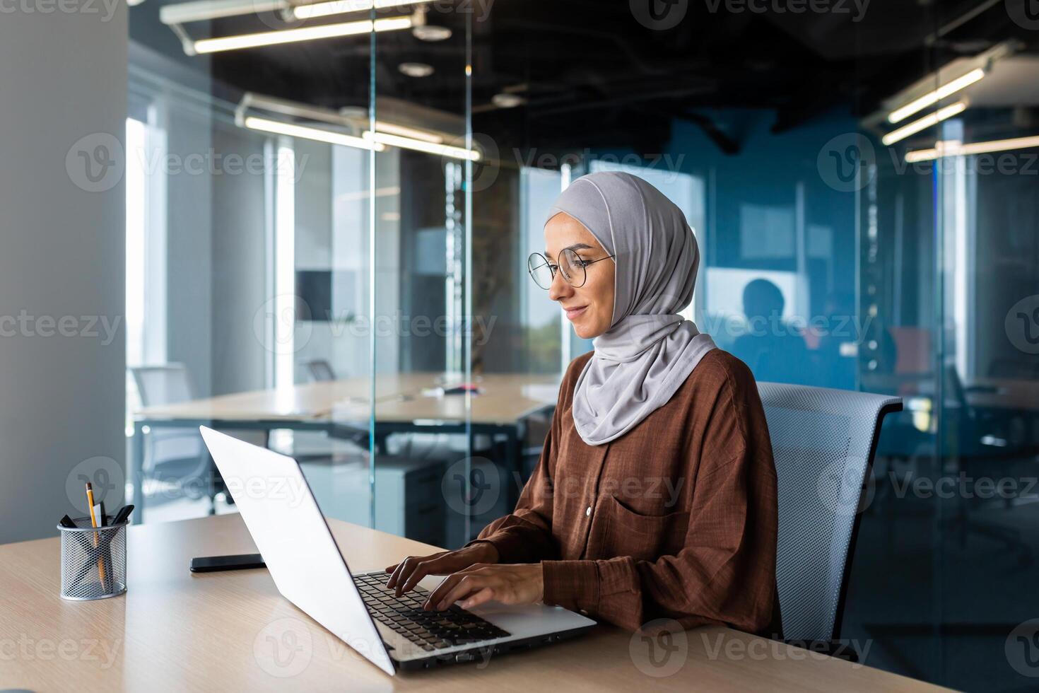Successful smiling Arab woman in hijab working inside modern office, Muslim woman using laptop at work, business woman satisfied with achievement results typing on computer keyboard. photo