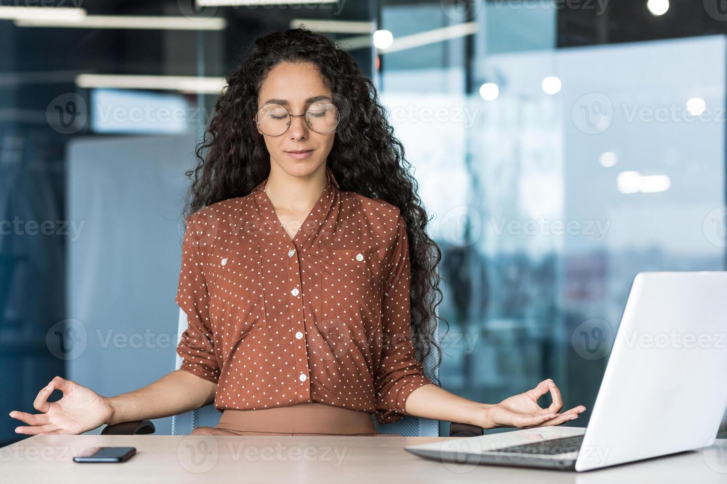 Young beautiful Hispanic web developer working inside office building, woman resting meditating at workplace with eyes closed, female worker using laptop to write code. photo