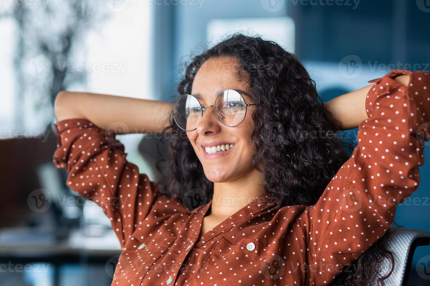 Close-up photo. Portrait of a young happy, successful Latin American woman. She is sitting in the office on a chair, hands behind her head, smiling, looking out the window. photo