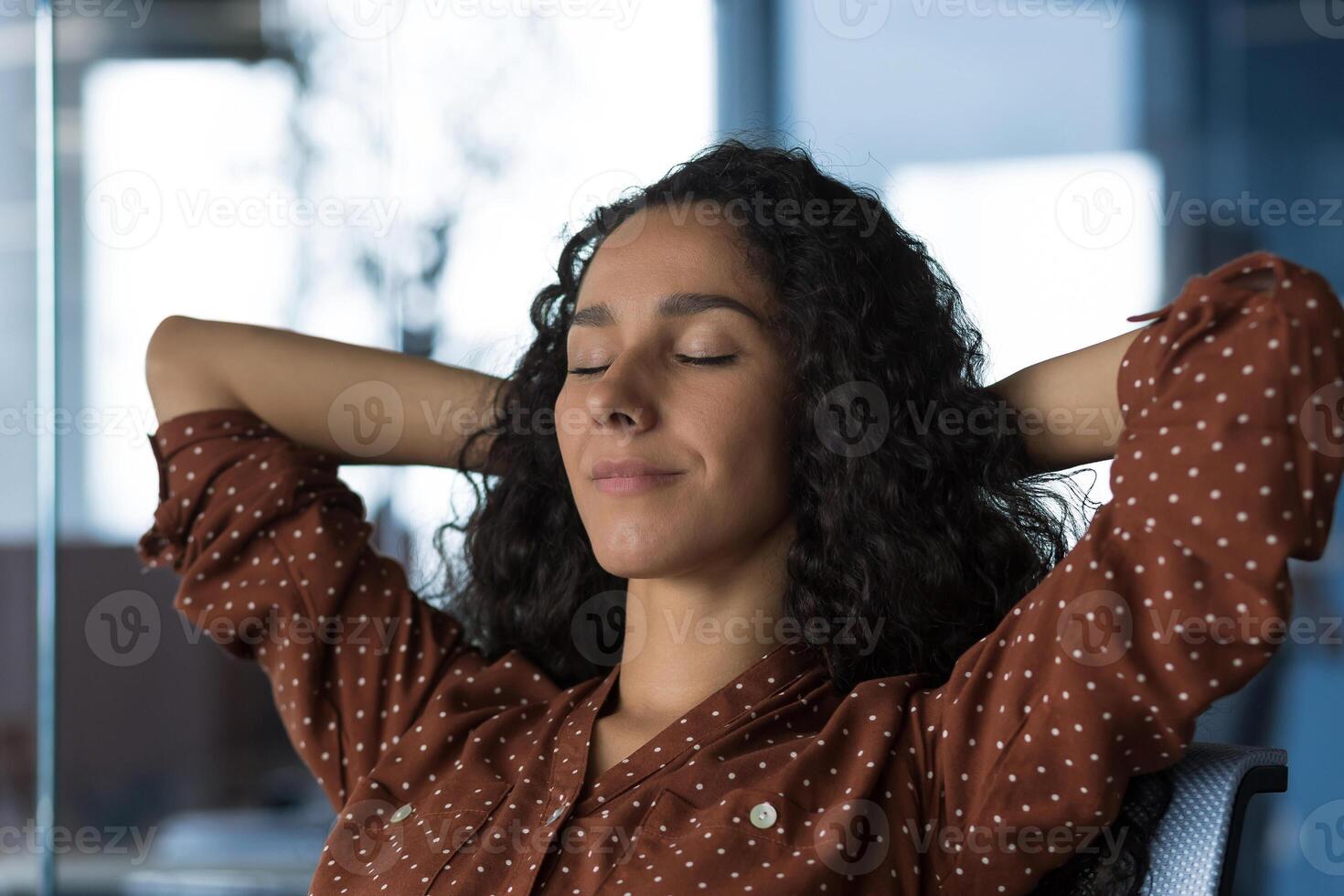 Close-up photo portrait of young beautiful curly Arab woman resting indoors at home, woman with hands behind head resting sitting on sofa near window, eyes closed breathing, meditating and dreaming