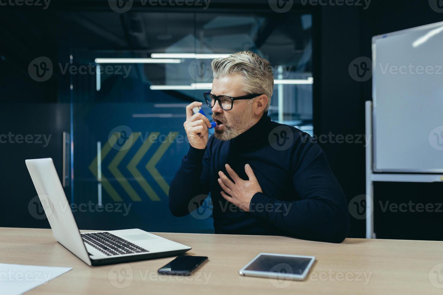 Senior gray-haired adult businessman sick at workplace inside office, man has asthma attack difficult to breathe, boss uses inhaler with medicine for relief. photo