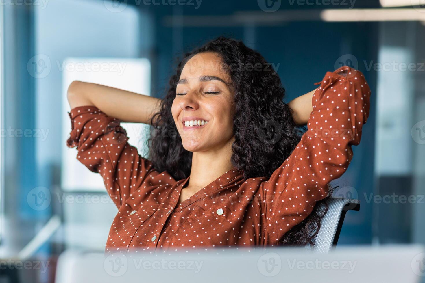 Close up pjoto. A young beautiful Latin American businesswoman sits in the office at a table with a laptop, hands behind her head, eyes closed, resting, smiling. photo