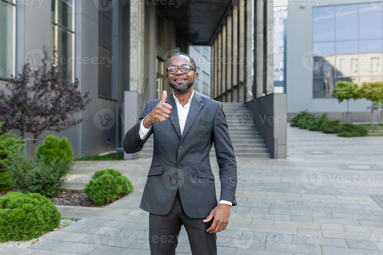 Portrait of a successful African American man. A businessman, a lawyer stands in front of an office center in a suit, looks at the camera, points a super finger, smiles. photo