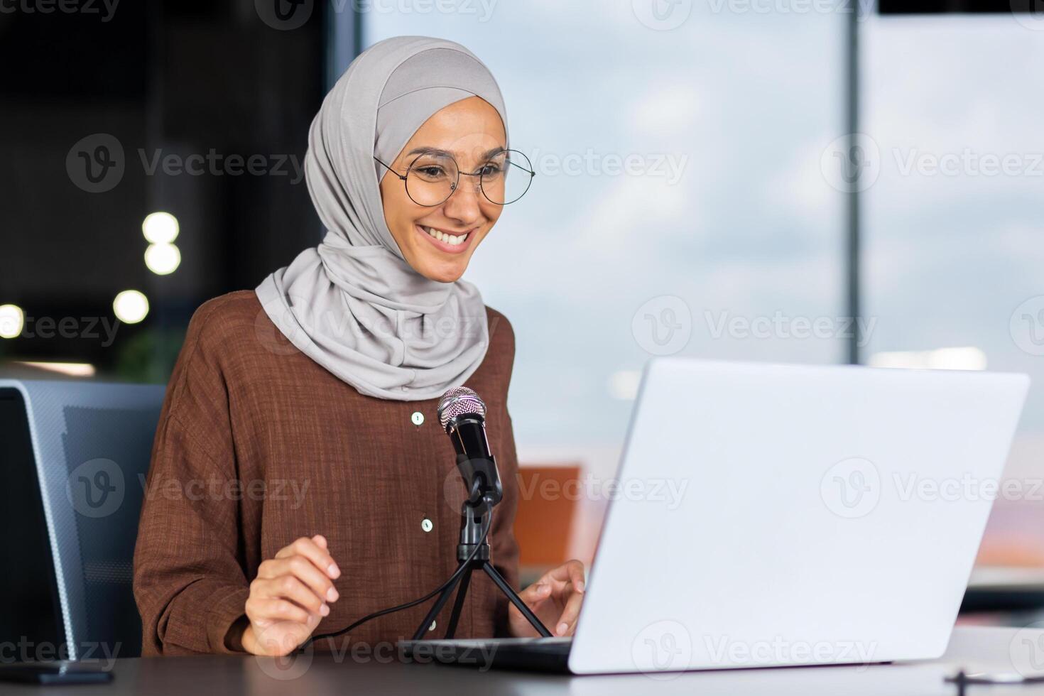 A young Muslim woman in a hijab is sitting in an office in front of a laptop and a microphone. records a podcast, a blog, talks on a call, gives an interview, goes through an online interview. photo