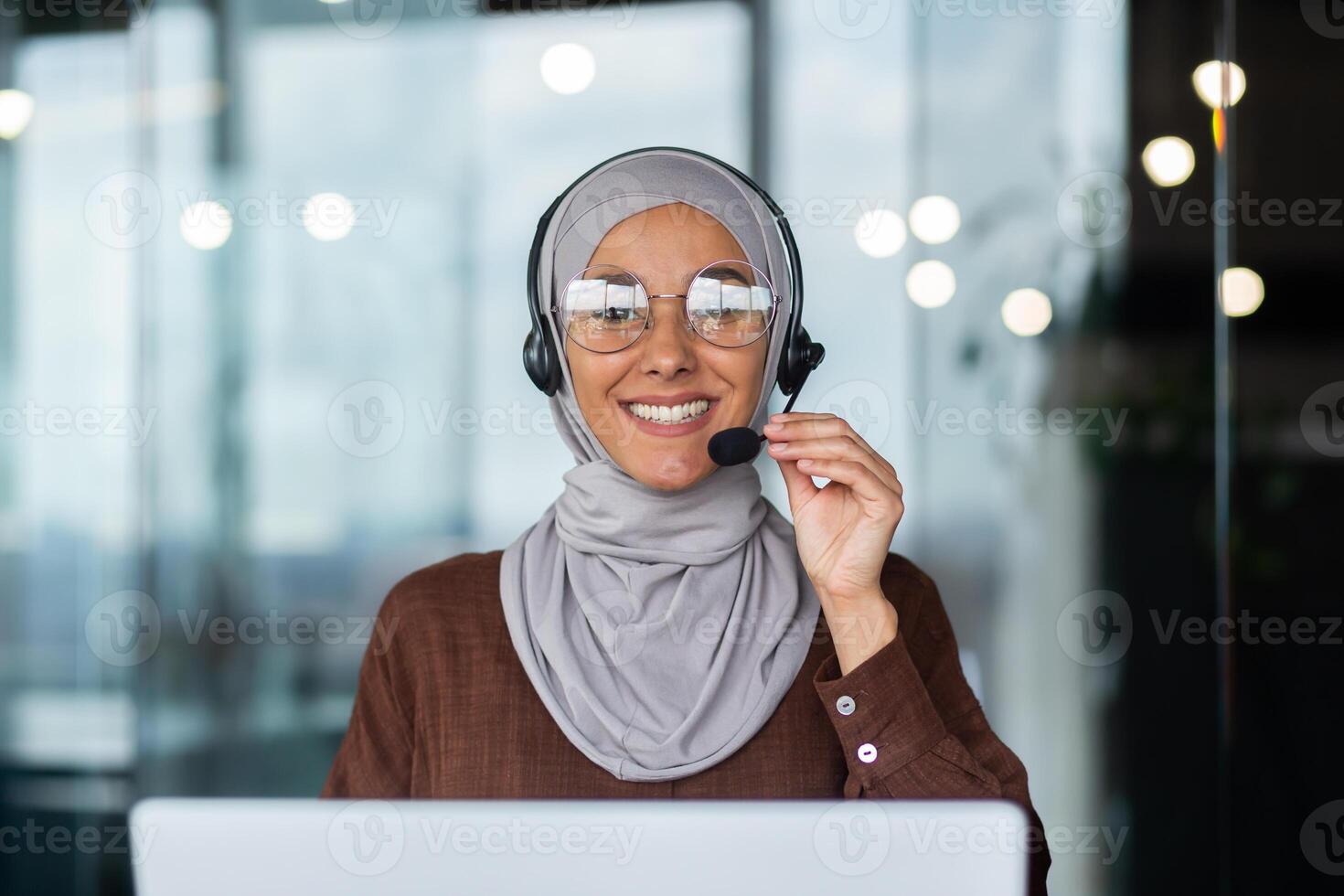 Close-up photo. Young hispanic woman in hijab sitting in office in headset and talking, consulting, advising on call from laptop. He holds the microphone in his hand, looks at the camera. photo
