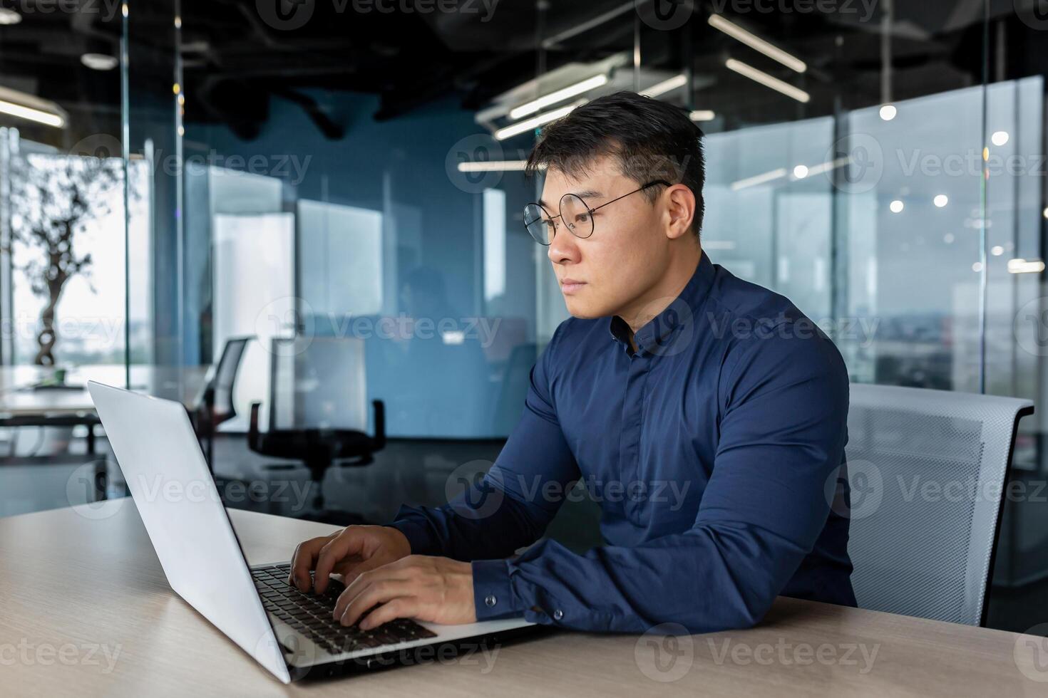 Serious and focused businessman working with laptop, man in glasses and shirt inside office building, asian boss typing on computer. photo