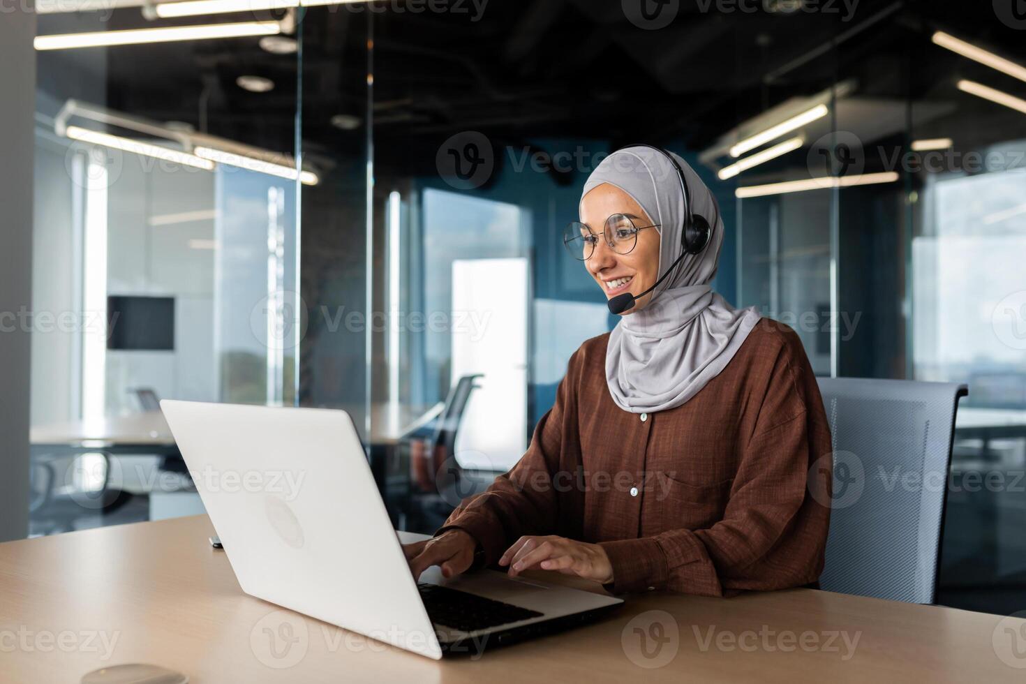 A young Muslim woman in a hijab and a headset is working in the office at a desk using a laptop. Typing on the keyboard, talking via call, consulting. photo