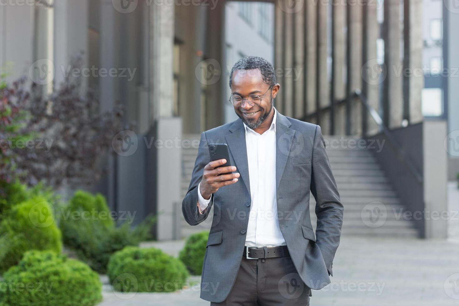 Successful african american businessman outside modern office building in business suit using smartphone, mature boss checking message smiling and reading news online from phone. photo