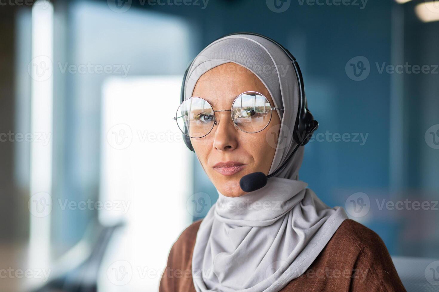 Close-up photo. Young Muslim woman in hijab and headset, helpline and call center consultant sitting in office at desk and looking seriously at camera. photo