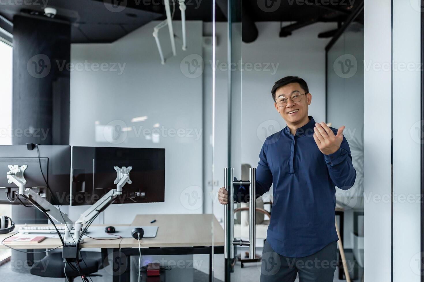 A successful engineer programmer boss invites to his office, an Asian man in a shirt and glasses looks at the camera and smiles and waves to the interlocutor. photo