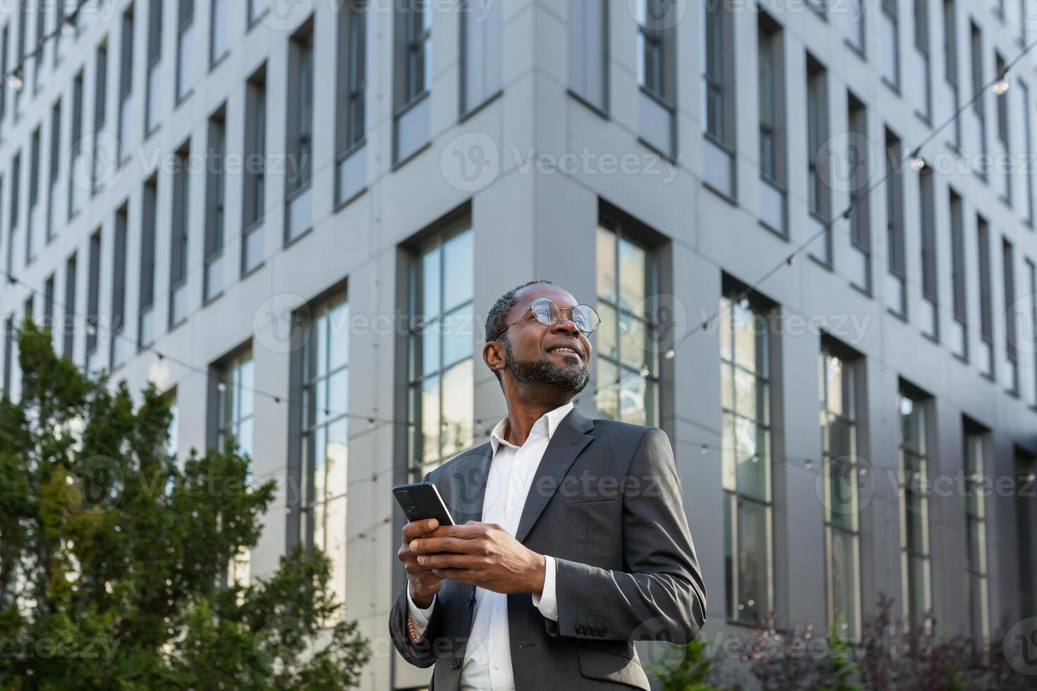 Successful african american boss businessman uses a telephone, man types messages and browses internet pages online on smartphone app outside office building walking in the city. photo
