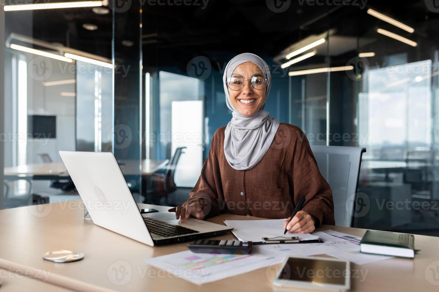 Portrait of young successful Arab woman in hijab working in office with documents and laptop. Smiling and looking at the camera. photo