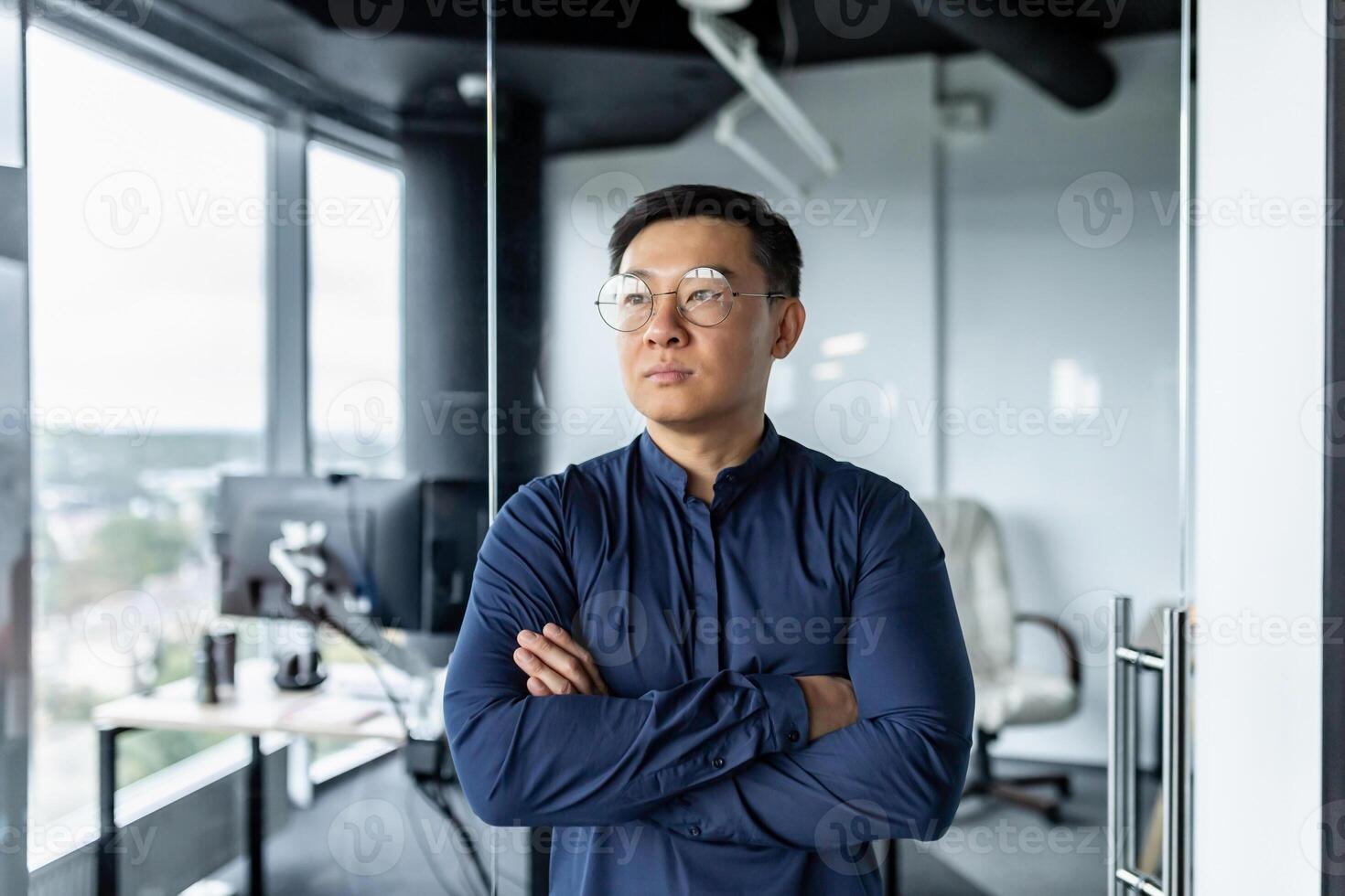 Thinking businessman with crossed arms in glasses looking out window, asian developer working inside modern office building. photo