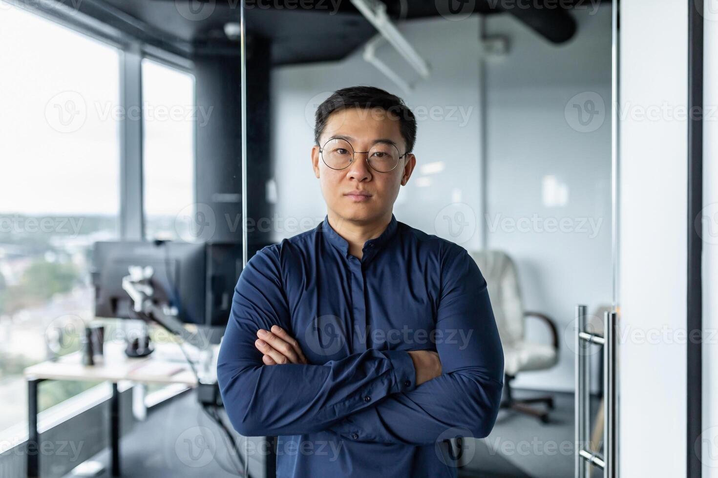 Portrait of serious Asian developer, businessman with crossed arms looking concentrated at camera, man in glasses and shirt working inside modern office building. photo