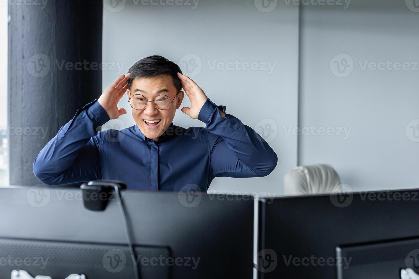 Happy business broker looking at two paired computer monitors and happy, businessman working inside modern office building, asian man celebrating successful deal and signed contract. photo