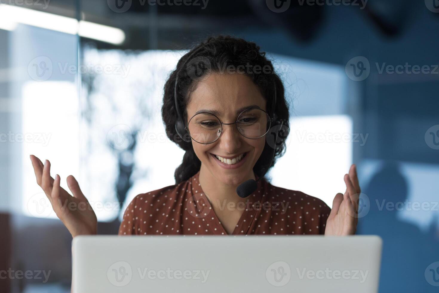 Close-up photo of young beautiful curly Arab woman talking on call, looking at laptop web camera and smiling using headset for communication, successful businesswoman working in office