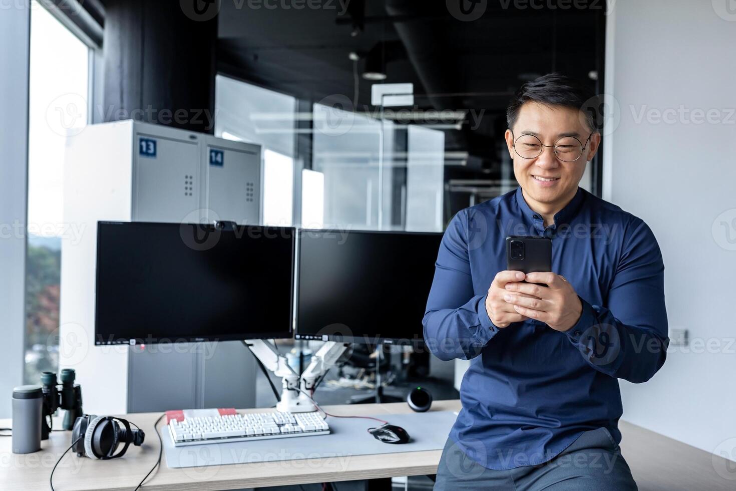Korean worker programmer developer working inside modern office building with dual computers, man using phone, typing message, Asian man reading good news online from smartphone. photo