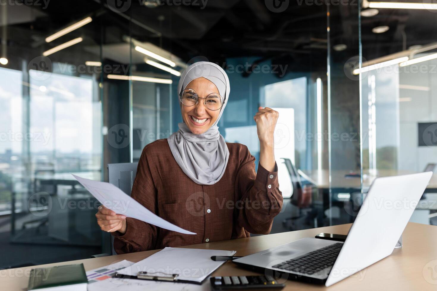 Young Muslim business woman in hijab is happy with success, profit and concluded deal. Sitting in the office at the table with documents and showing a victory gesture with her hand to the camera. photo