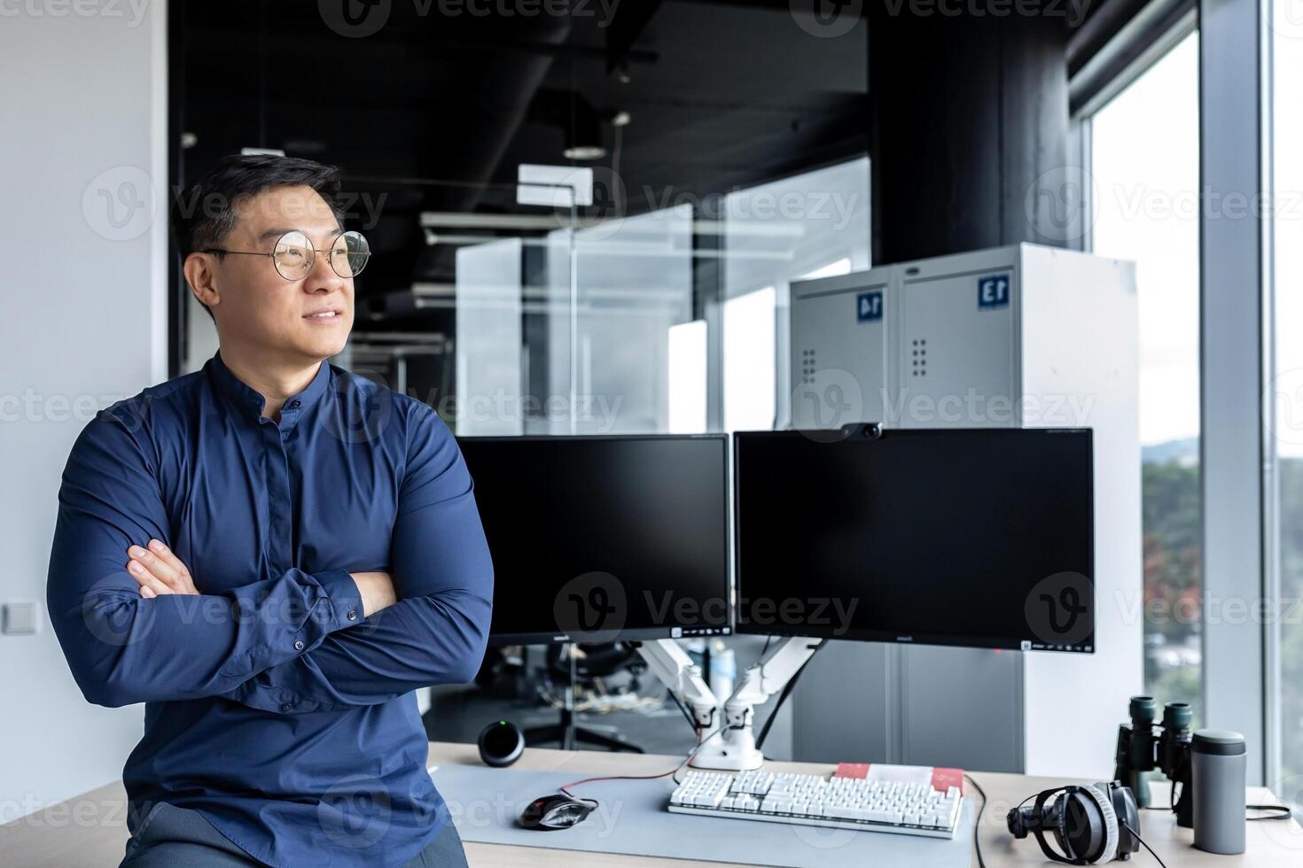 Portrait of Asian businessman, man in glasses and shirt looking out window and smiling, worker inside office building with crossed arms. photo