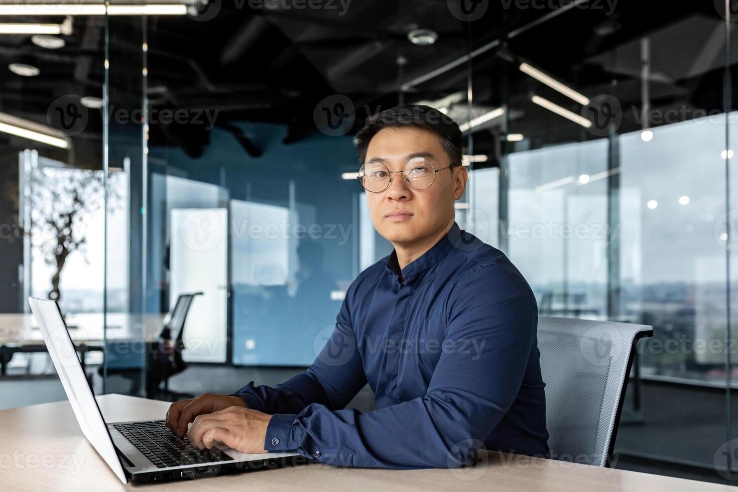 Serious and focused businessman looking at camera, asian man working inside modern building, man wearing glasses and casual shirt using laptop. photo