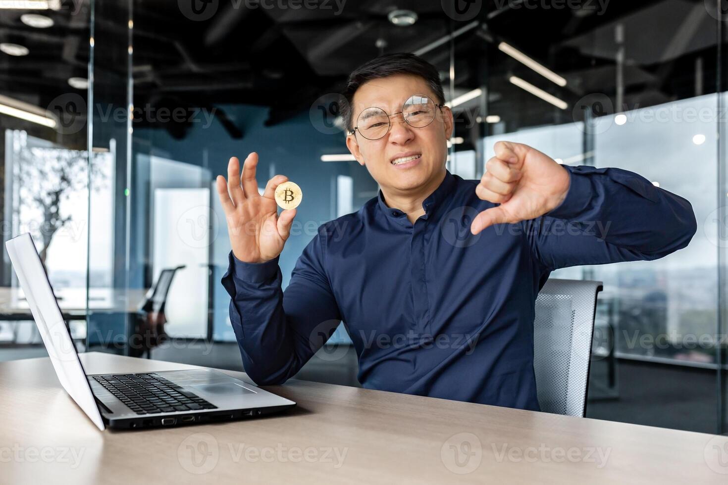 Dissatisfied asian businessman holding finger down, bad losing gesture, holding virtual currency bitcoin money, man looking disappointed at camera, working inside modern office building. photo