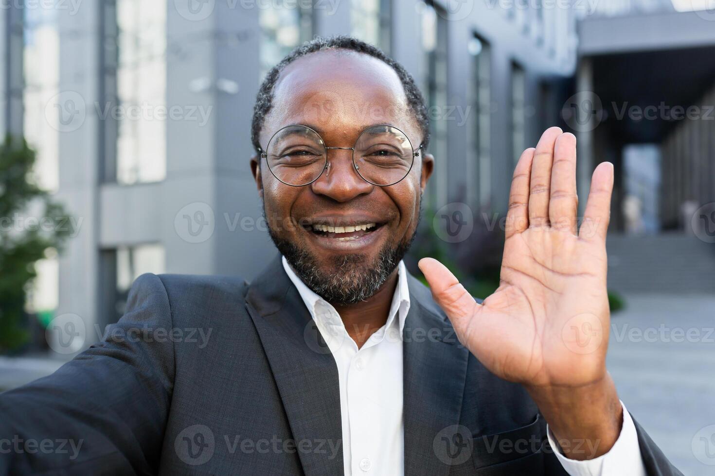 Close-up photo. An African American businessman in a suit and glasses stands outside an office center and talks on a call. He is holding a phone, waving at the camera, smiling. photo
