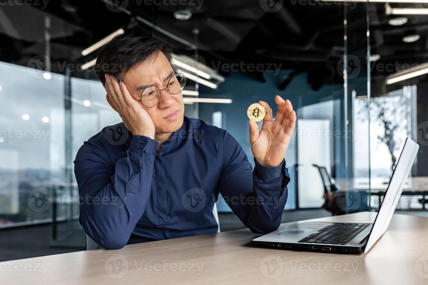 Frustrated asian businessman investor holding crypto currency bitcoin in hands, man working inside modern office building, failure with financial investment. photo