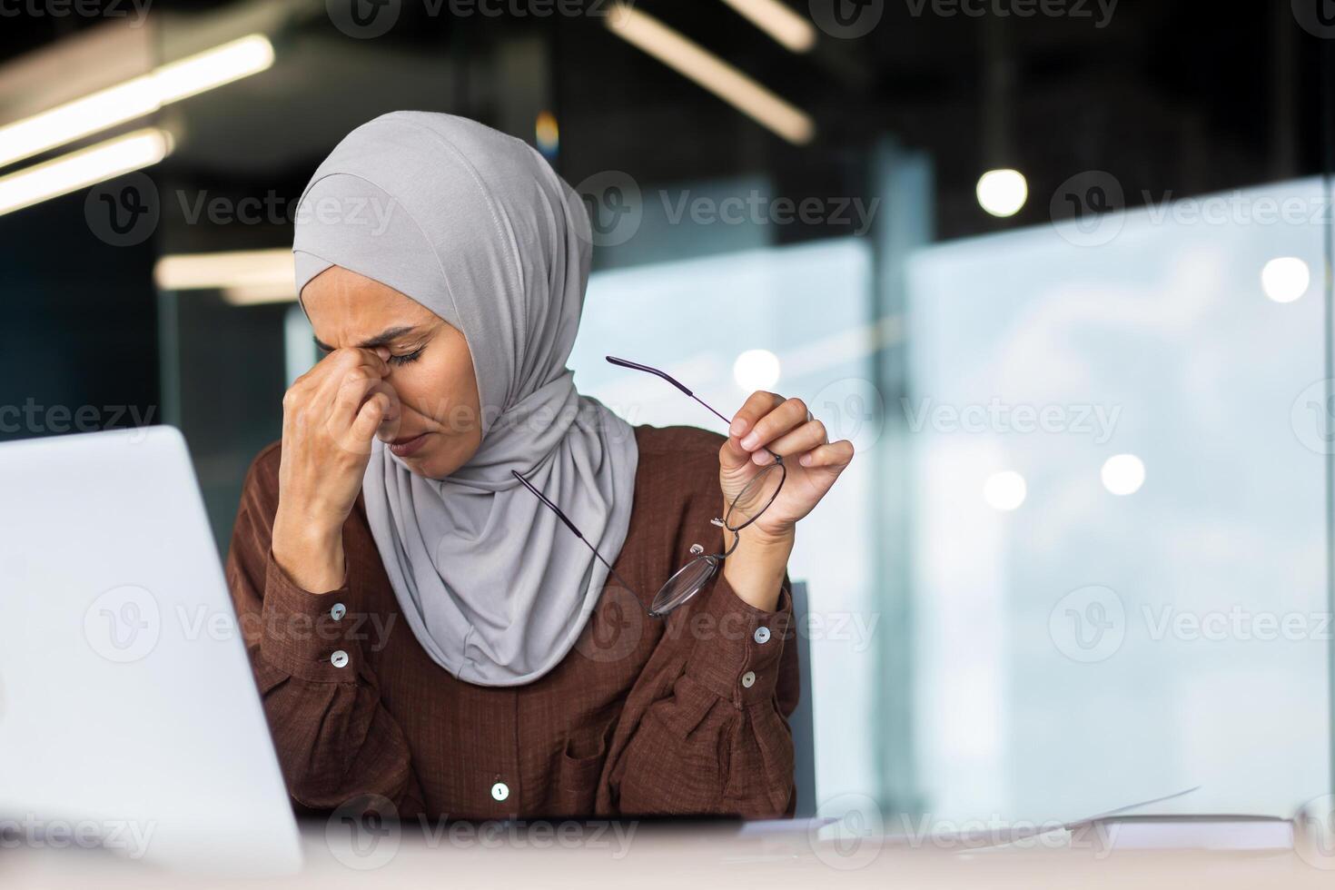 Tired young Muslim female teacher in hijab sits at desk in front of laptop. She holds glasses in her hand, closed her eyes, rubs her eyes with her hand, rests, takes a break in online learning. photo