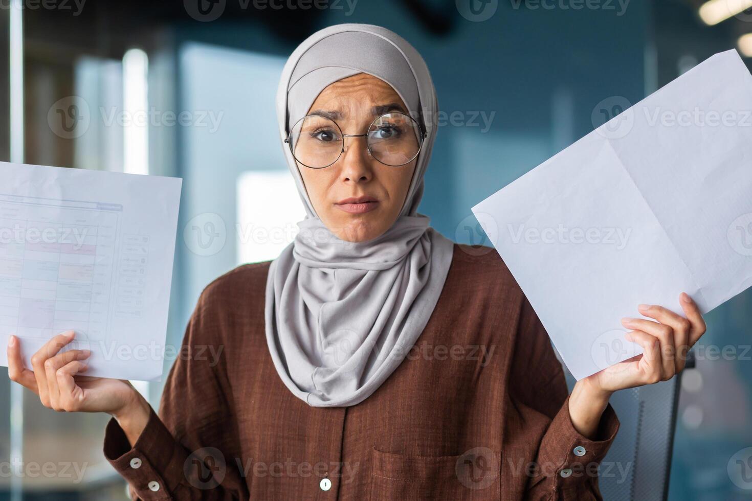 Burned out at work, financial problems. Portrait of a young hijabi Muslim woman sitting in the office, looking worriedly at the camera and holding documents, charts, a project in her hands. photo