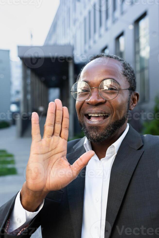 Close up photo. Smiling african american man in suit and glasses standing near office center talking on call. He waves his hand at the camera, greeting. photo