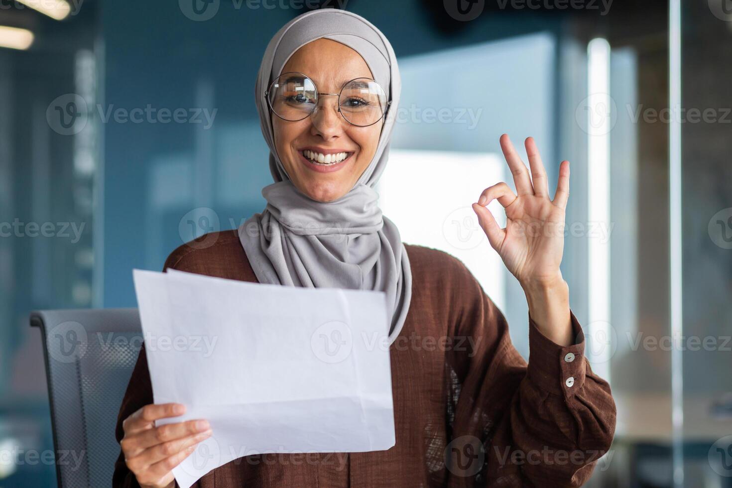 Portrait of a happy Muslim woman in a hijab holding a document, a letter, a sheet of paper. Got good news, new job, promotion. He smiles and points to the camera with his hand ok. photo