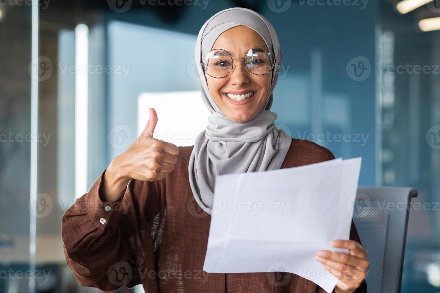 Portrait of a happy Muslim woman in a hijab, a student holding a document, a letter, a sheet of paper in her hands. Received good news, smiles and points to the camera with a super hand. photo