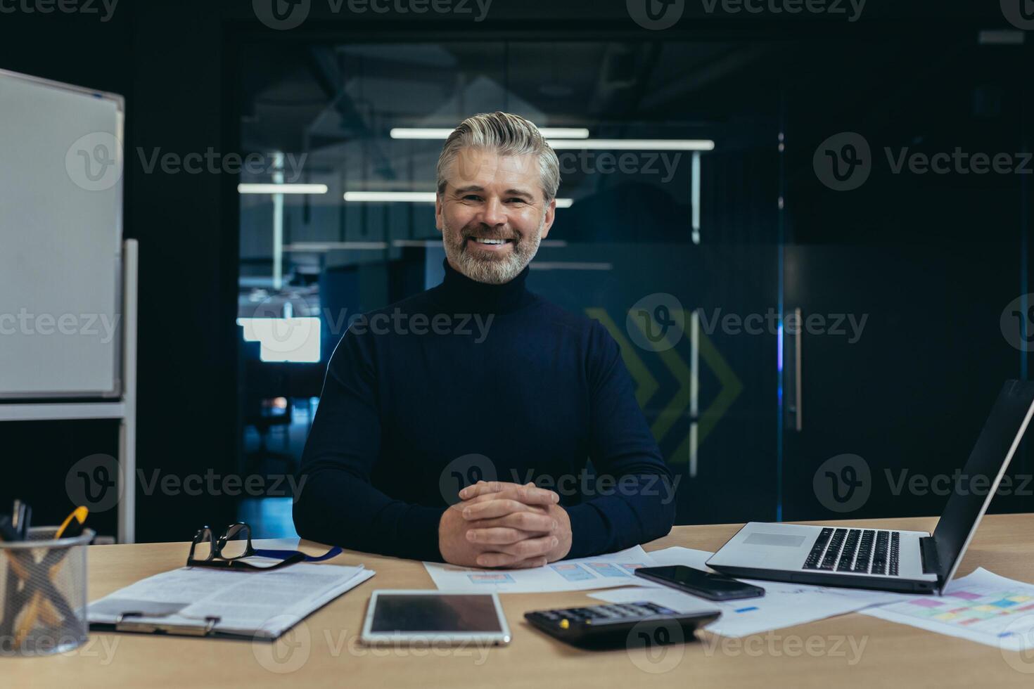 Portrait of a self-confident successful senior man businessman, accountant, manager sitting in the office at the table, smiling and looking at the camera, hands folded together. photo