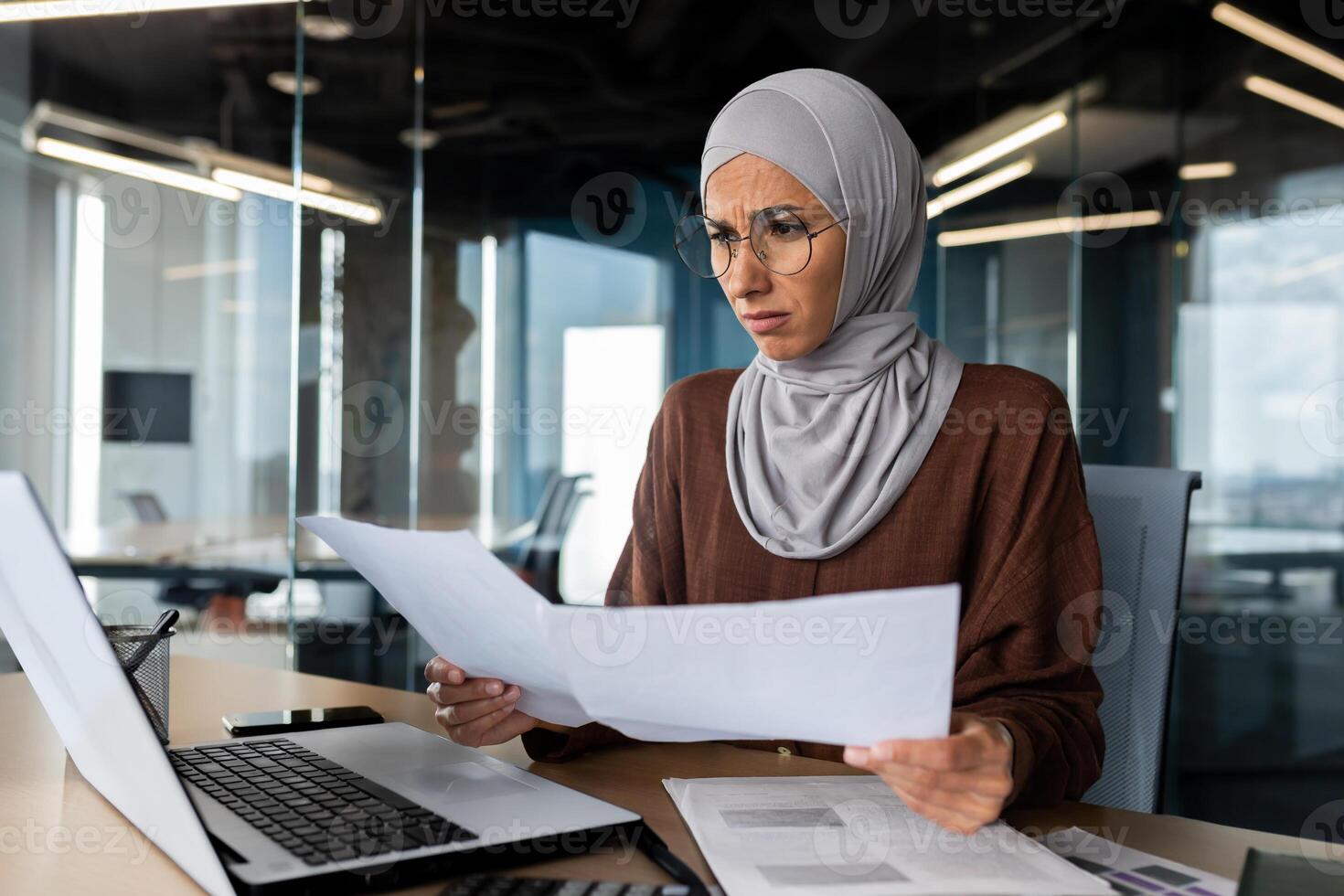 Burned out at work, financial problems. Confused young hijabi Muslim woman sitting in office, working on laptop and holding documents, graphs, project in hands. photo