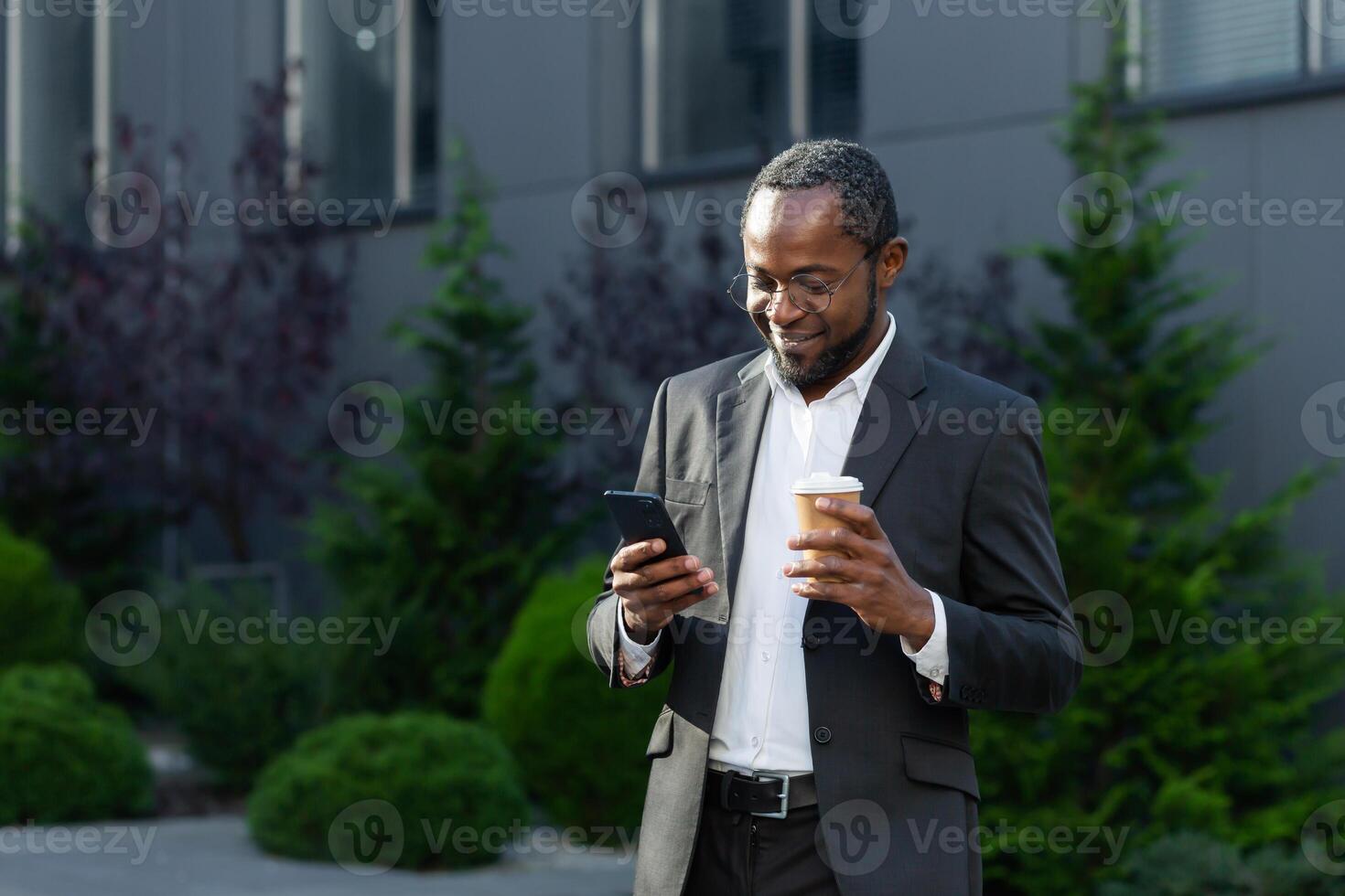 Mature businessman in business suit using phone outside office building, african american boss holding cup of hot drink, smiling typing text message and browsing websites. photo