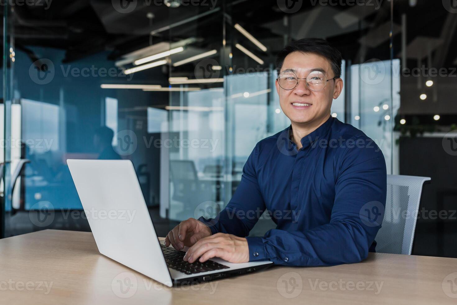 Young and successful asian businessman in glasses working on laptop inside modern office building, happy manager smiling and looking at camera, entrepreneur investor portrait. photo