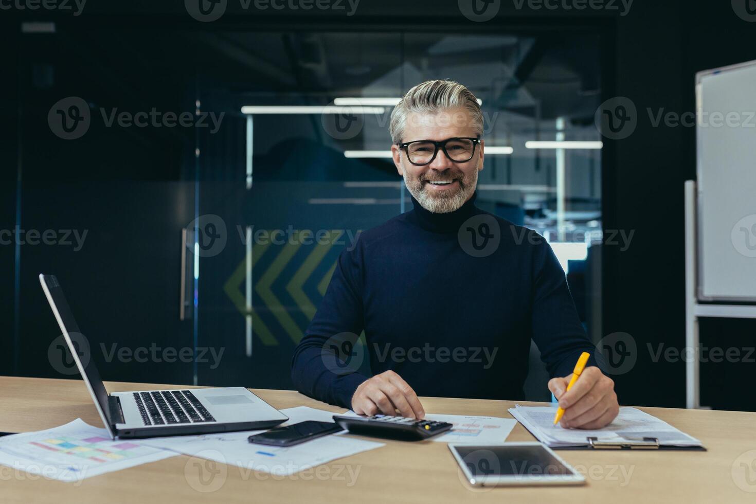 Portrait of an older gray-haired man, an accountant, an auditor sitting in the office at a laptop and a calculator. He works with documents, checks accounts. Smiling at the camera. photo