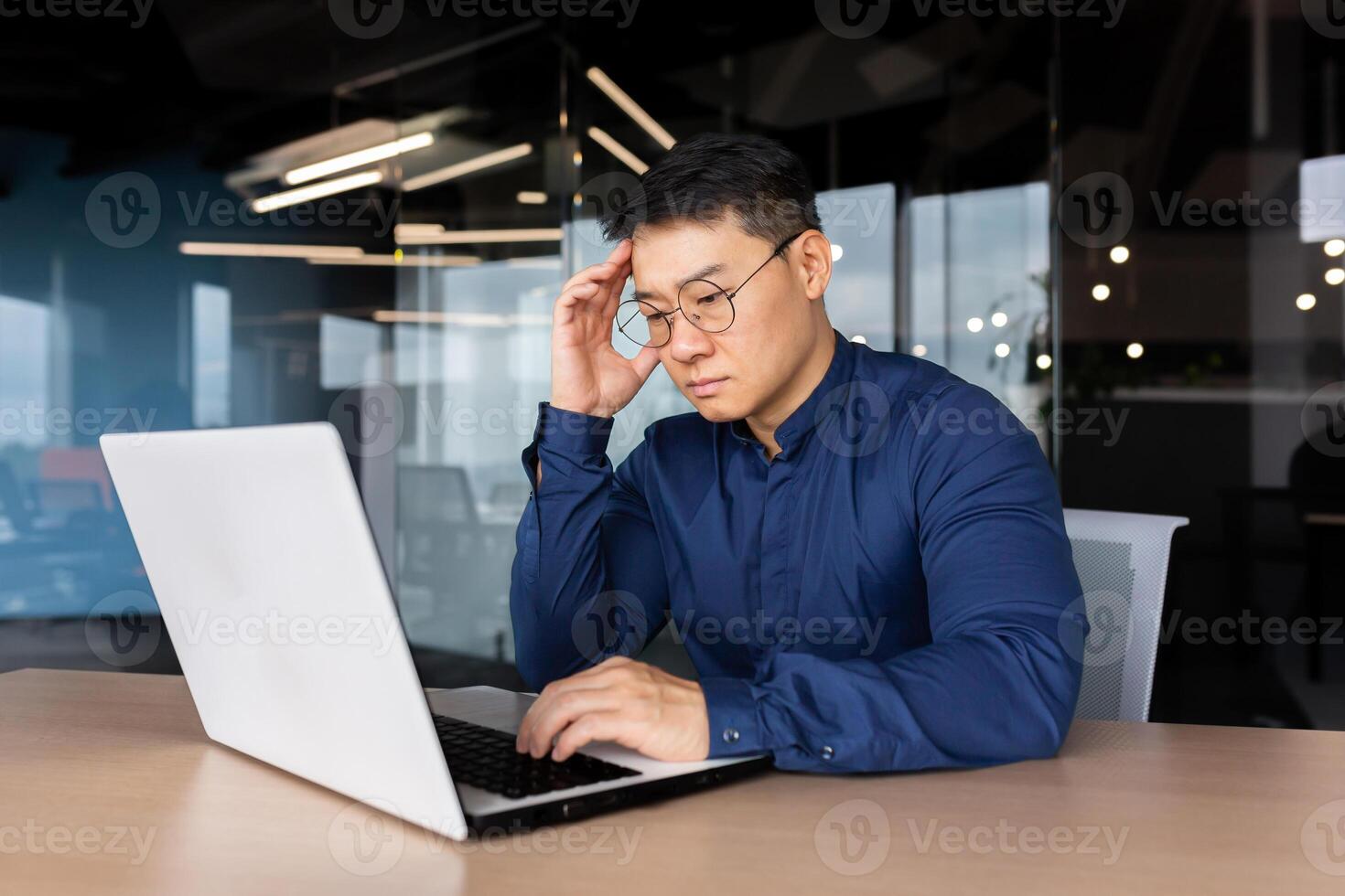Thoughtful and serious male Asian student sits in the office at the table, works on a laptop. Passes the exam, worries, studies online through a call. He holds his head with his hand. photo