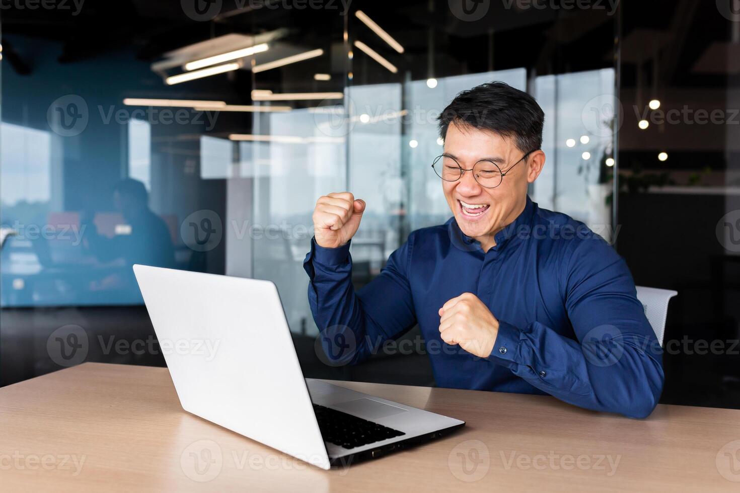 A young male Asian student received good news, mail, passed the exam, entered the university, received a scholarship. He sits in the office, looks at the monitor, rejoices, shows yes with his hand photo