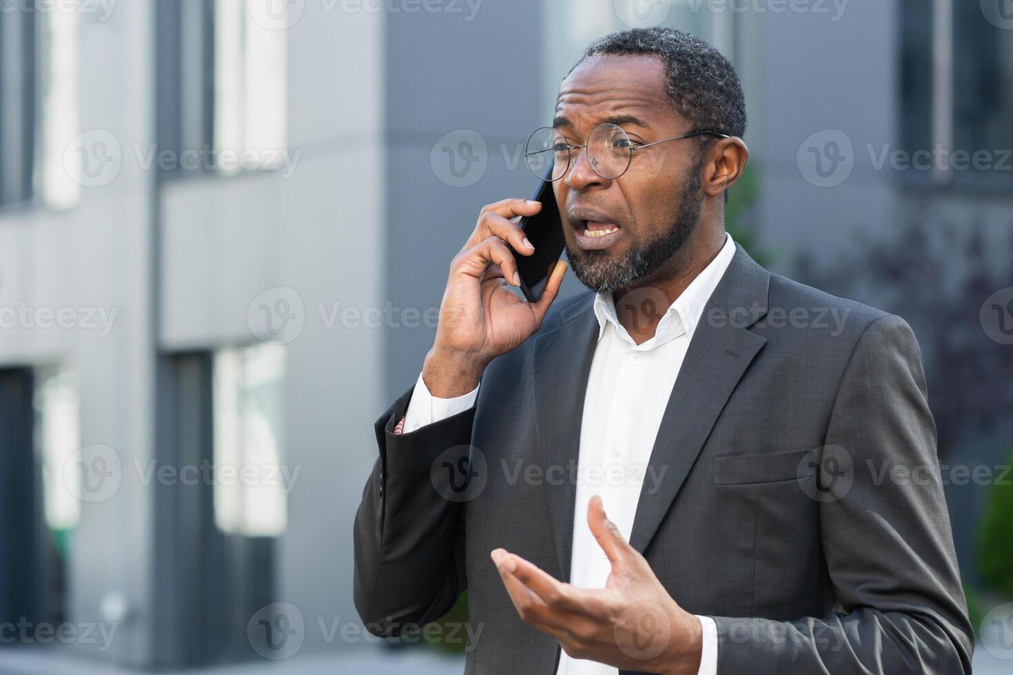 Angry and angry boss talking and shouting on mobile phone, african american businessman in business suit walking outside office and talking with subordinates. photo