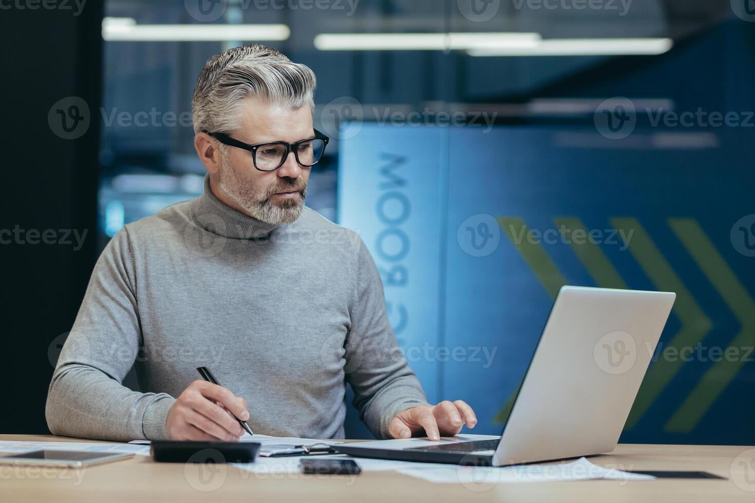 serious businessman behind paper work in office, gray-haired investor signing documents, man working in modern office with accounts and laptop, gray-haired and mature financier photo