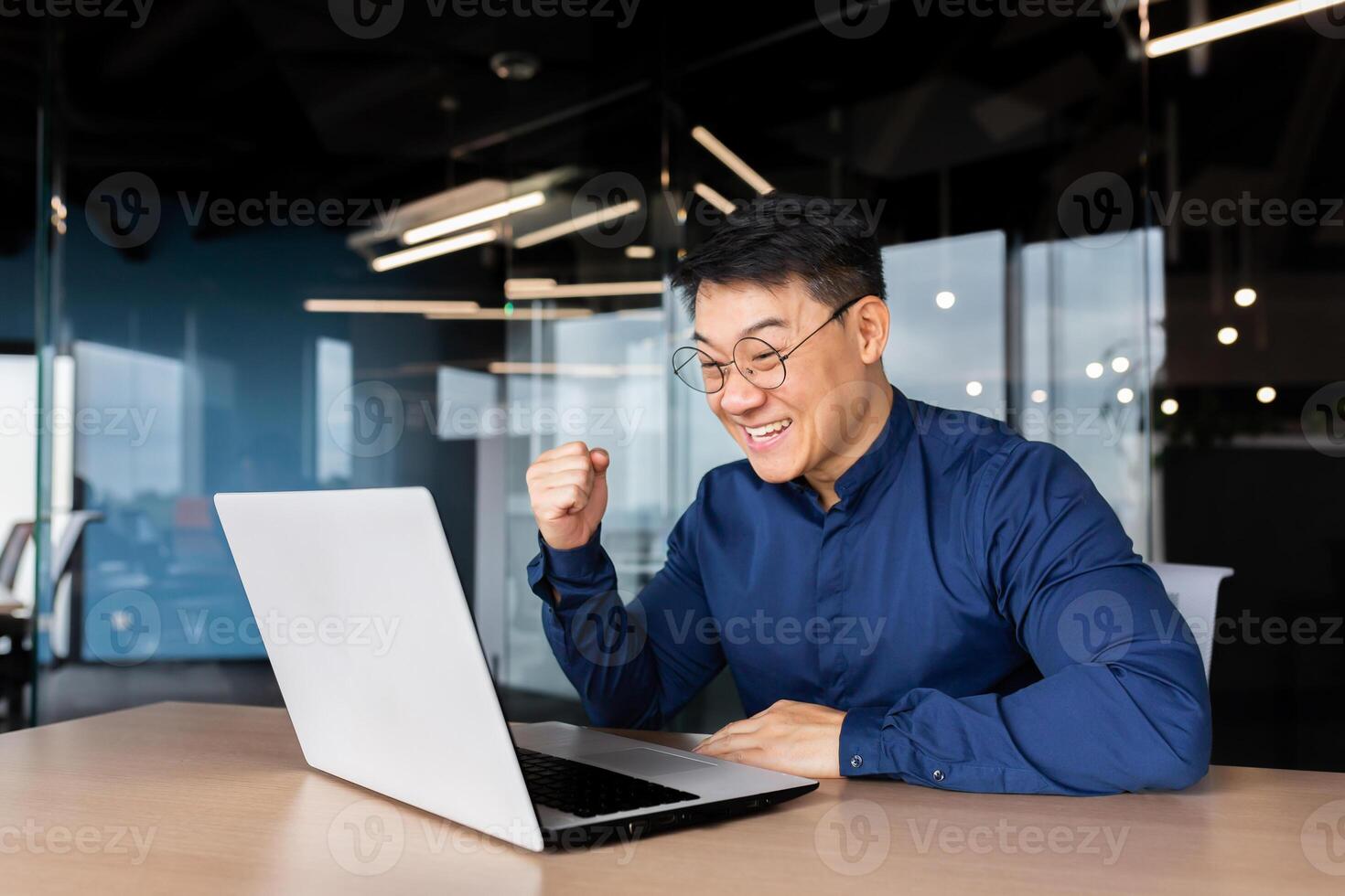 A happy young Asian man sits in the office at the table, looks at the laptop, talks on a call, online meeting, rejoices at the success, the deal, the win. photo