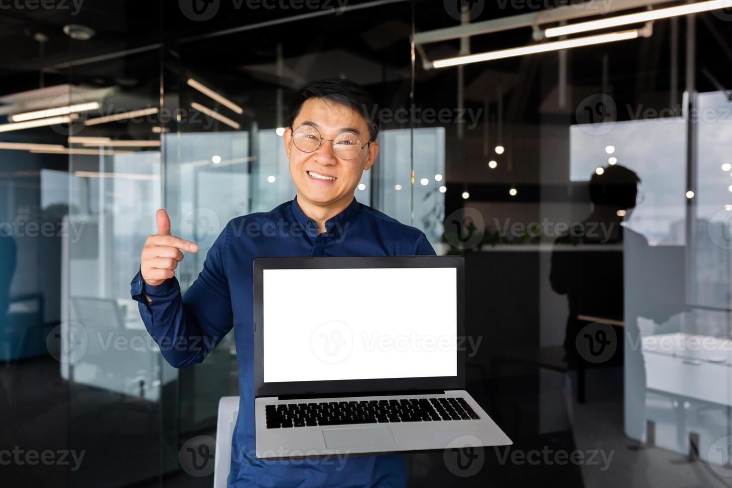 Portrait of smiling successful asian man inside office, man smiling and looking at camera standing holding laptop and pointing finger at white screen, businessman in shirt and glasses at workplace. photo