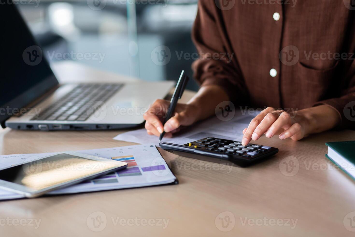 Close-up photo. Hands of a young woman in a brown shirt calculating on a calculator and writing documents at an office table. photo