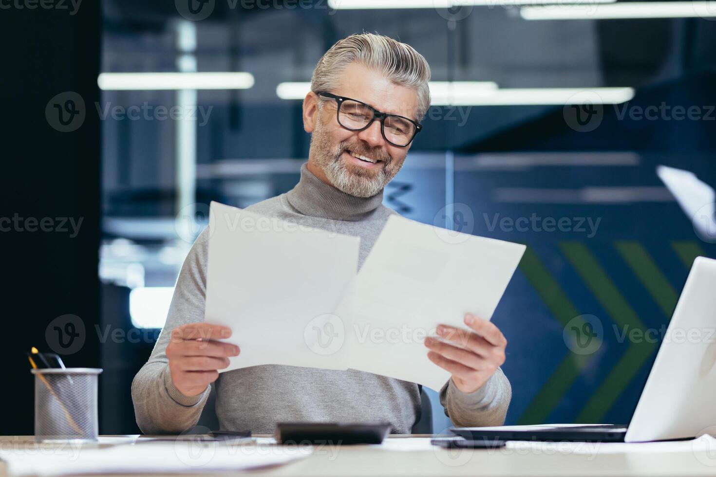 Senior smiling gray-haired businessman working in office at desk with laptop and documents. Holds in his hands, looks at sheets of paper, agreements, bills. photo