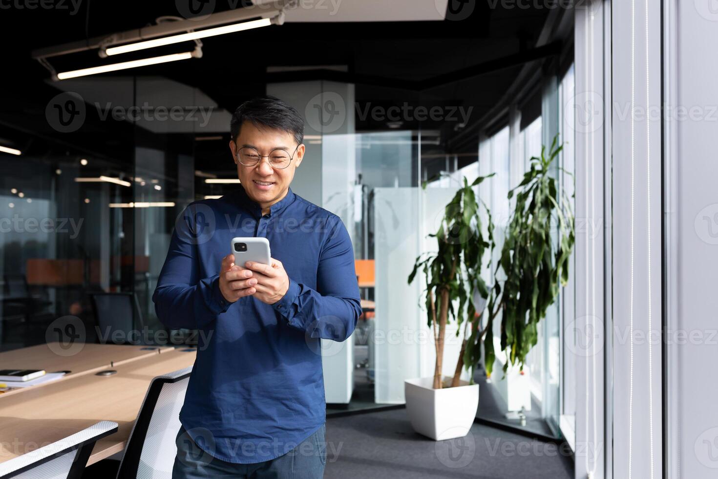 A young man, an Asian businessman, a student standing by the window in the office, uses a mobile phone, types messages, reads, checks mail. He looks at the camera, smiles. photo