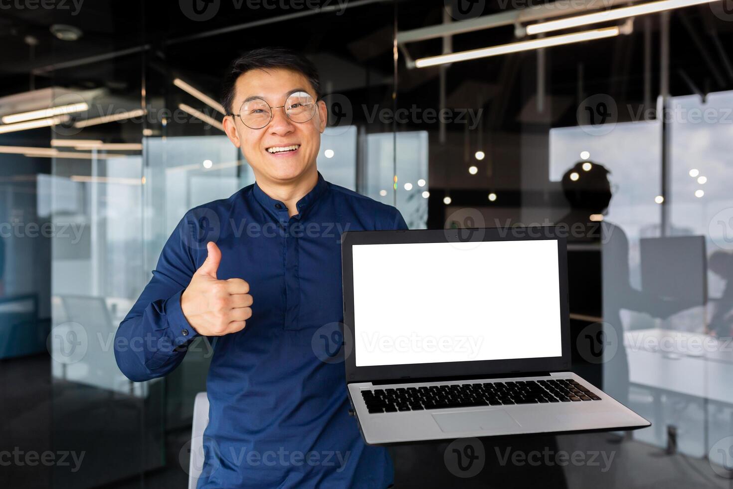 An Asian man inside the office smiles and looks at the camera, a businessman holds a laptop with a blank screen in his hands, an investor shows a thumb up and positively recommends cooperation. photo